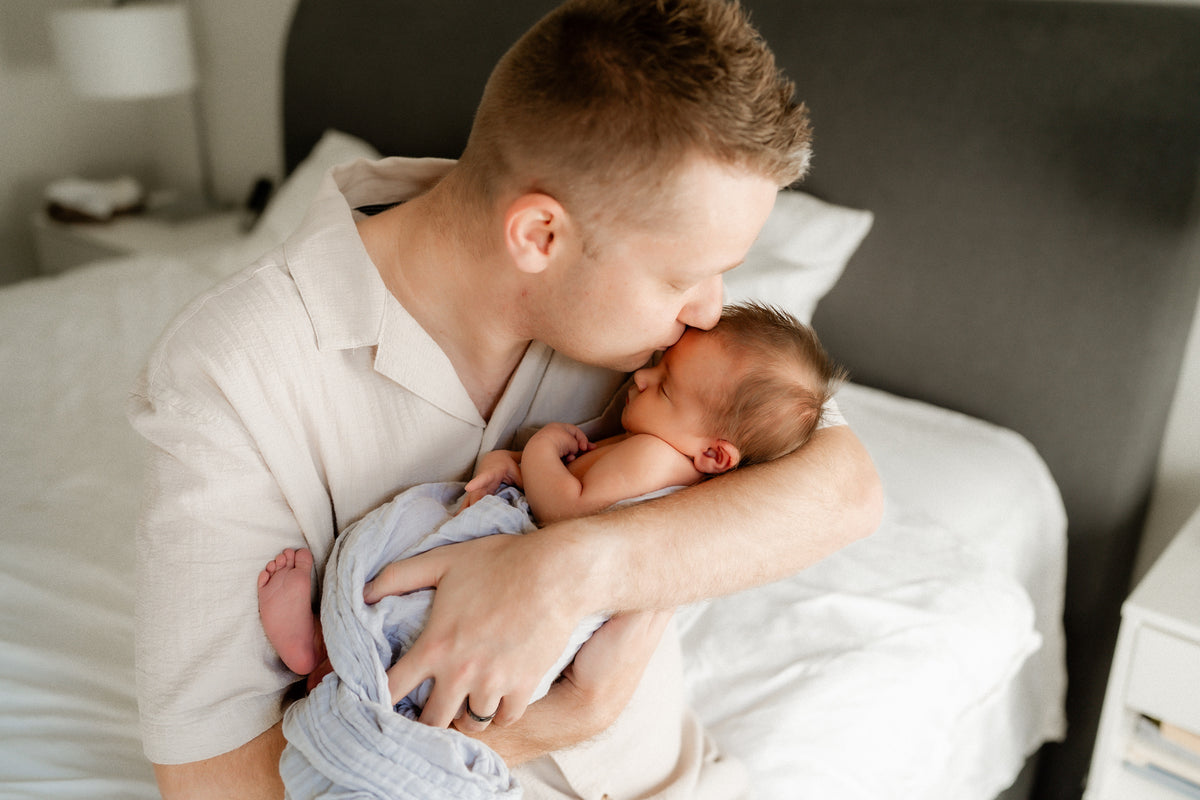 A man in a light beige linen shirt holds and kisses a sleeping newborn baby wrapped in a light blue blanket, sitting on a bed with white linens and a dark gray headboard.
